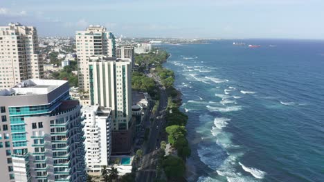 Panoramic-aerial-view-of-skyscrapers,-buildings-and-blue-waves-of-the-Caribbean-sea-in-the-center-of-Malecon,-waves-breaking-on-the-coast,-Dominican-Republic
