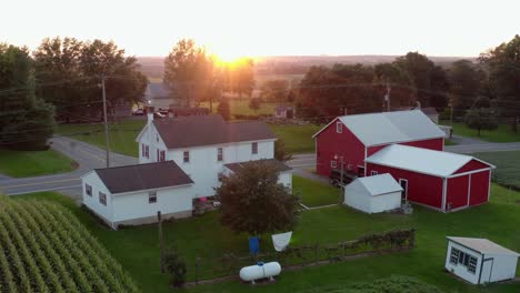 Aerial-establishing-shot-of-white-house,-red-barn-in-rural-America,-USA-at-summer-sunrise,-sunset