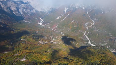 Mist-over-the-alpine-valley-with-riverbed-across-mountain-slopes-covered-in-colorful-forest-trees-in-Autumn