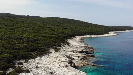 Moving-Along-the-Rocky-Shoreline-Covered-with-Trees-and-Shrubs-Besides-Clear-and-Calm-Turquoise-Blue-Waters-of-Paralia-Emplisi-Beach-in-Erisos,-Greece---Forward-Moving-Aerial-Shot
