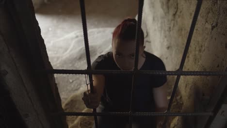 A-young-woman-kneeling-in-a-cell-clutches-the-bars-and-raises-her-head-to-stare-in-a-terrifying-way
