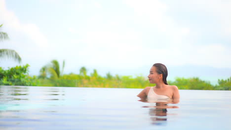 Tropical-island-resort-luxury-destination-with-attractive-Asian-lady-relaxing-in-calm-swimming-pool-overlooking-the-lush-landscape-and-ocean-in-background