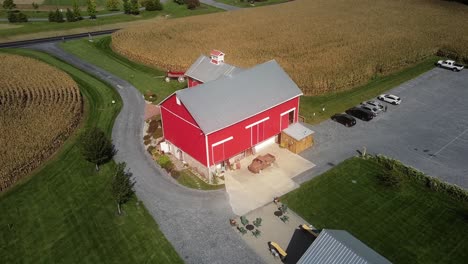 Aerial-drone-view-of-a-red-barn-wedding-venue-with-garden-party-concept,-red-house-flanked-by-wheat-fields-and-driveway-seen-from-the-road,-wedding-hall