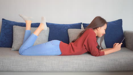 Young-woman-laying-on-stomach-using-smartphone-in-living-room-on-sofa-checking-online-profile-or-dating-app