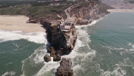 Waves-crashing-on-rocky-cliffs-with-Fortress-of-Saint-Michael-the-Archangel-and-Nazare-lighthouse-suited-on-top
