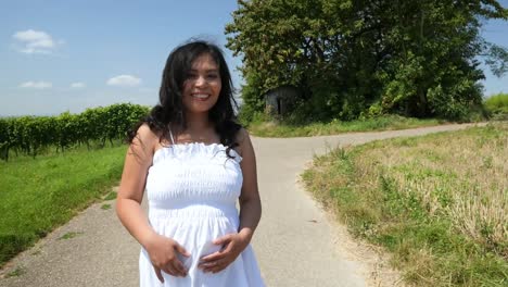 Junge-Schwangere-Frau-In-Einem-Weißen-Sommerkleid,-Die-Eine-Landstraße-Hinuntergeht-Und-Ihren-Bauch-Für-Ein-Mutterschaftsfoto-Shooting-Hält