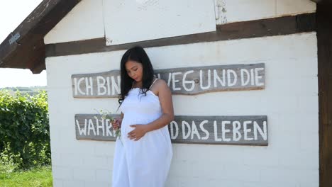 Young-pregnant-Woman-in-a-White-Summer-Dress-leaning-against-a-wall-with-Flowers-in-her-Hands-for-a-Maternity-Photo-Shoot