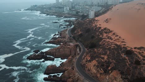 Aerial-dolly-in-of-coastal-road-and-orange-sand-dunes-in-rocky-hillside-near-sea-waves-revealing-Concon-buildings-on-a-foggy-day,-Chile