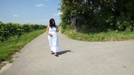 Young-pregnant-Woman-in-a-White-Summer-Dress-walking-down-a-Country-Road-holding-her-belly-and-flowers-for-a-Maternity-Photo-Shoot