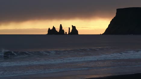 Wide-shot-Silhouette-of-Reynisdrangar-basalt-sea-stacks-beside-mountain-during-sunset-light-in-Iceland---Waves-reaching-beach-during-mysterious-sky---Rock-formation-in-background