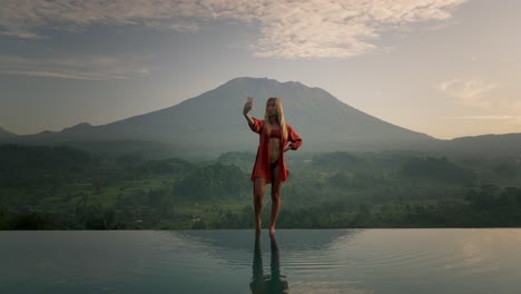 Modelo-Rubia-Posando-En-El-Borde-De-La-Piscina-Infinita-En-Bikini-Durante-El-Amanecer,-Bali-Mount-Agung
