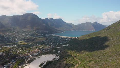Overlooking-the-Harbor-Town-of-Hout-Bay-Surrounded-By-Mountains-and-the-Hout-Bay-Beach-in-the-Distance-in-Cape-Town,-South-Africa---Wide-Angle-Aerial-Shot