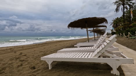 4k-aufnahme-Eines-Leeren-Strandes-An-Einem-Bewölkten-Tag-Mit-Sonnenliegen-Und-Strohschirmen-In-Marbella,-Spanien