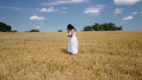 Young-pregnant-Woman-in-a-White-Summer-Dress-standing-on-a-field-holding-her-belly-and-flowers-for-a-Maternity-Photo-Shoot---Drone-Circle-Close