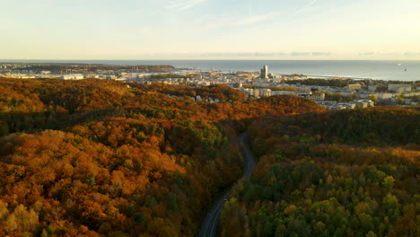 Aerial---Car-Driving-Between-Hills-Through-the-Colorful-Autumn-Forest-At-Sunset-with-Gdynia-city-Skyline-and-Baltic-Sea-Shore-on-background---pull-back-shot