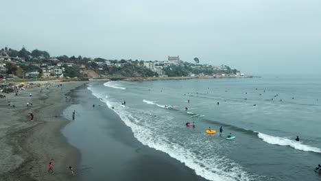 Aerial-orbit-of-people-swimming-and-surfing-in-the-sea-near-the-sand-shore-in-La-Boca-beach,-Concon-hillside-neighborhood-in-background,-Chile