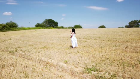 Young-pregnant-Woman-in-a-White-Summer-Dress-standing-on-a-field-holding-her-belly-and-flowers-for-a-Maternity-Photo-Shoot---Drone-Circle-Far