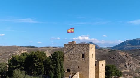 Antequera-Alcazaba-Schloss-Mit-Spanischer-Flagge,-Die-Mit-Bergen-Im-Hintergrund-Weht