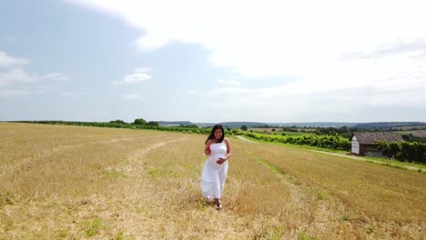 Young-pregnant-Woman-in-a-White-Summer-Dress-walking-down-a-field-holding-her-belly-and-flowers-for-a-Maternity-Photo-Shoot