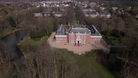 Sideways-aerial-pan-around-Slot-Zeist-castle-with-the-moated-manor-surrounded-by-green-park-and-urban-landscape-in-the-background