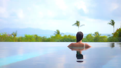A-woman-with-her-back-to-the-camera-looks-out-from-a-resort-pool-to-the-tropical-paradise-and-the-ocean-beyond