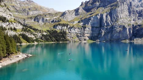 Aerial-flyover-over-the-turquoise-water-of-lake-Oeschinensee-in-Kandersteg,-Switzerland-with-views-of-boats-and-cliffs-on-a-sunny-summer-afternoon