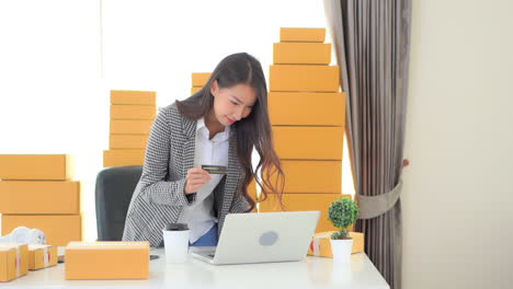 Asian-Woman-Received-Big-Payment-on-card-balance-doing-an-online-sales-with-her-laptop-computer,-courier-parcel-boxes-at-the-home-office
