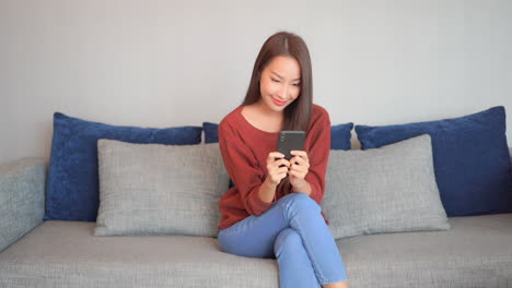 A-young-woman-sits-on-a-huge-couch-backed-by-decorative-pillows-as-she-focuses-intensely-on-the-input-to-her-smartphone