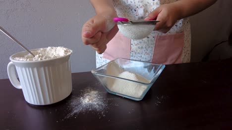 Latin-woman-wearing-an-apron-preparing-cooking-baking-a-cake-sieving-the-flour-using-her-hand-and-a-metal-strainer