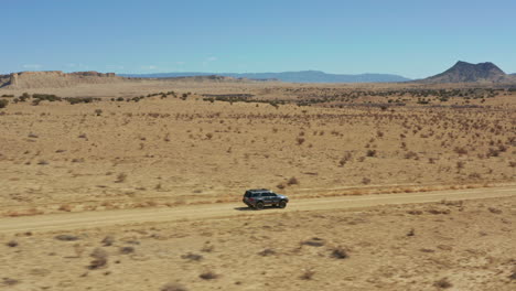 Aerial-following-SUV-down-dirt-road-with-wide-open-desert-landscape-behind