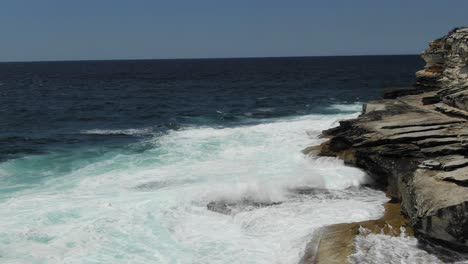 Drone-flying-along-rocky-coast-with-ocean-waves-breaking-on-cliff-near-Bondi-Beach,-Australia