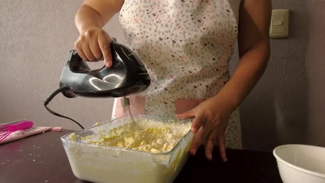 Lateinische-Frau,-Die-Eine-Schürze-Trägt,-Die-Das-Kochen-Vorbereitet,-Einen-Kuchen-Backend,-Der-Alle-Zutaten-In-Einem-Behälter-Mit-Dem-Mixer-Mischt