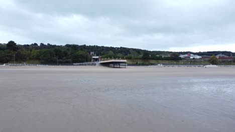 Colwyn-Bay-Welsh-seaside-town-pier-boardwalk-aerial-view-over-moody-overcast-sandy-low-tide-beach-flying-forwards