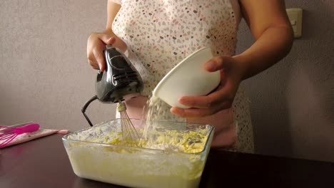 Lateinische-Frau,-Die-Eine-Schürze-Trägt,-Die-Das-Kochen-Vorbereitet,-Einen-Kuchen-Backend,-Der-Mehl-In-Die-Buttermischung-Gießt