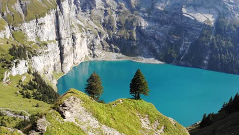 Aerial-flyover-over-trees-overlooking-the-turquoise-lake-Oeschinensee-in-Kandersteg,-Switzerland-surrounded-by-cliffs