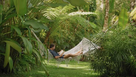 Young-man-lying-in-hammock-reading-book-in-jungle-paradise-surrounded-by-plants