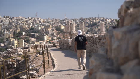 Man-Walking-by-Walls-of-Ancient-Amman-Citadel,-Jordan-With-Ruins-and-Cityscape-in-Background,-Slow-Motion,-Full-Frame