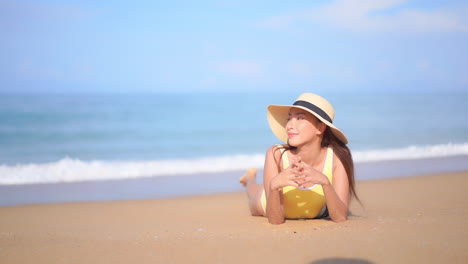 Woman-yellow-swimsuit-and-sunhat-resting-lying-down-leaning-on-elbows-at-a-sandy-beach-near-the-sea-when-foamy-waves-roll-over-the-sand