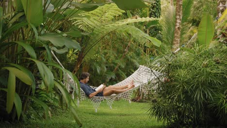 Young-man-lying-in-hammock-reading-a-book-in-green-jungle-paradise-surrounded-by-plants