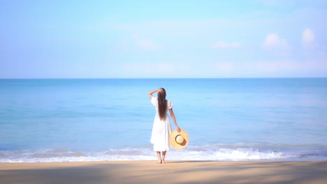 Asian-Woman-standing-on-the-beach-by-the-sea-and-raising-hands-up-wearing-white-summer-dress-and-holding-straw-sunhat-in-hand---vacation-template-back-view-copy-space