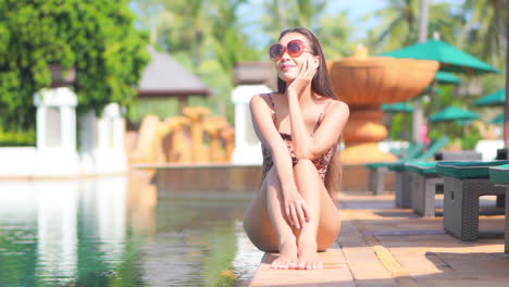 Young-adult-Asian-woman-in-swimsuit-and-sunglasses-sitting-at-the-edge-of-swimming-pool-with-tropical-greenery-background-next-to-line-on-deckchairs-in-Thailand-resort-slow-motion
