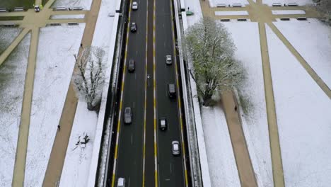 Traffic-flows-through-patterned-public-sidewalks,-newly-fallen-snow-covers-the-grass,-aerial