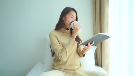 Junge-Asiatische-Attraktive-Frau,-Die-Ein-Interessantes-Buch-Liest-Und-Heißen-Tee-Oder-Kaffee-Aus-Einer-Weißen-Tasse-Auf-Der-Couch-Im-Gemütlichen-Wohnzimmer-Trinkt