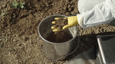 Soil-sampling-concept,-field-ground-in-gloved-hands-and-bucket,-high-angle-closeup