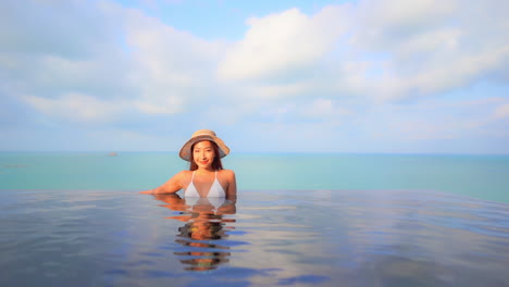 Mujer-Asiática-En-La-Piscina-Infinita-De-Un-Complejo-Hotelero-De-Lujo-Mirando-A-La-Cámara-Con-La-Luz-Del-Sol-En-La-Cara,-Cuadro-Completo