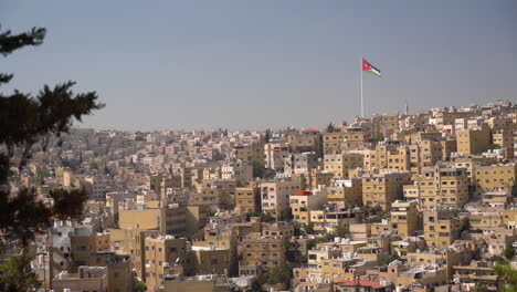 Jordanian-Flag-Waving-on-Hilltop-Above-Hillside-Residential-Neighborhood,-Amman,-Jordan-on-Suny-Day