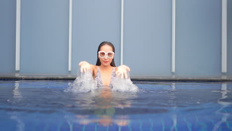 Asiatische-Frau-Im-Schwimmbad,-Die-Eine-Weiße-Sonnenbrille-Trägt,-Die-Wasser-Mit-Beiden-Händen-Spritzt