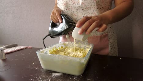 Mujer-Latina-Con-Un-Delantal-Preparando-La-Cocina-Horneando-Un-Pastel-Vertiendo-Leche-En-La-Masa-Y-Usando-La-Batidora