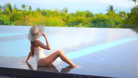 Petite-Exotic-Woman-in-Swimsuit-and-Floppy-Hat-at-Luxury-Poolside-With-Stunning-View-on-Tropical-Vegetation-and-Sea,-Full-Frame