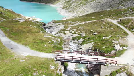 Vuelo-Cercano-Sobre-Un-Pequeño-Puente-De-Montaña-Que-Revela-Un-Lago-De-Montaña-Azul-Turquesa-En-Un-Paisaje-Montañoso-En-Los-Alpes-Suizos